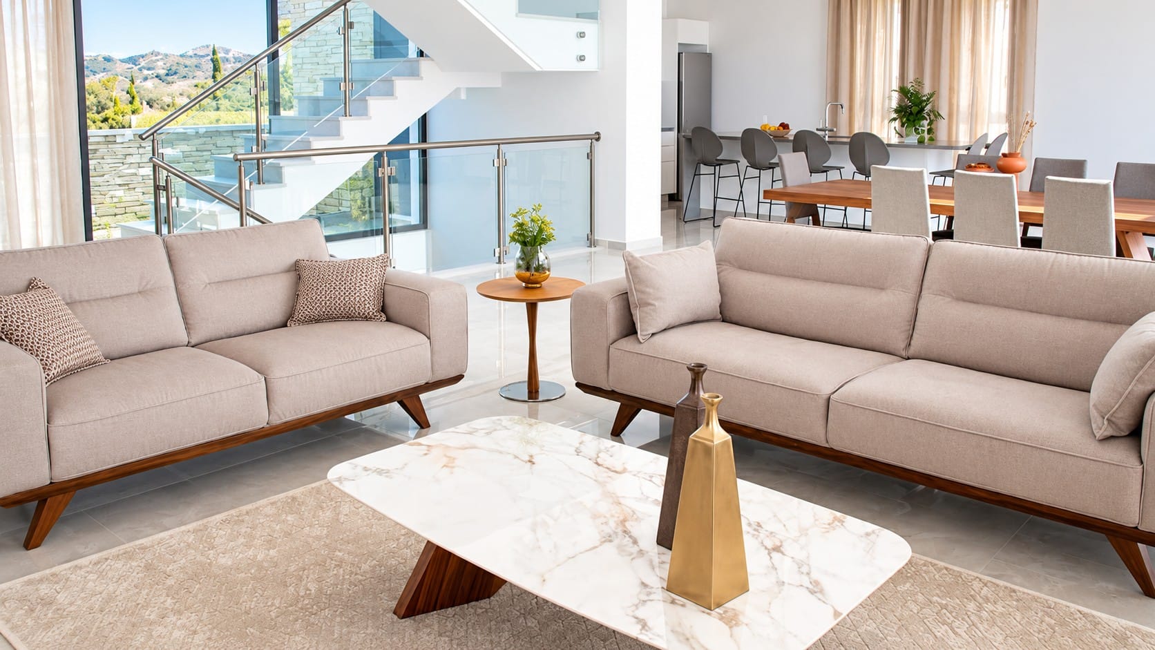 Living area with sofas, glass-balustrade staircase, dining table and kitchen breakfast bar, with hillside views through full-height windows