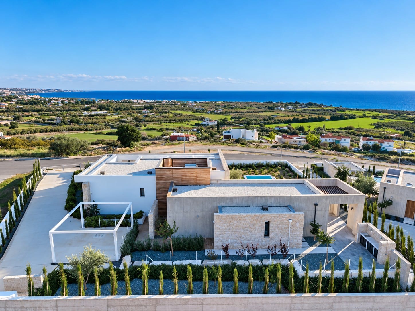 Aerial view of a single-level villa with flat roofs, pergola, stone and timber-clad facades and landscaped grounds, overlooking the Mediterranean Sea