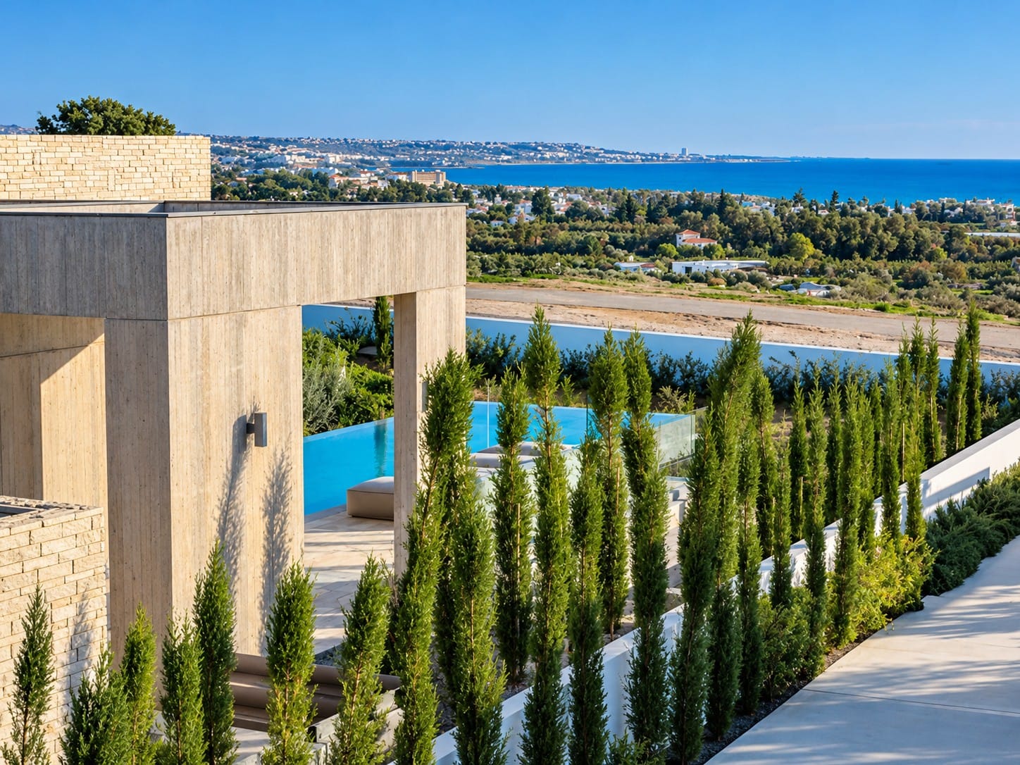 Villa exterior with stone and textured facades, rows of cypress trees and a glimpse of the swimming pool, with views towards the coastline and Mediterranean Sea