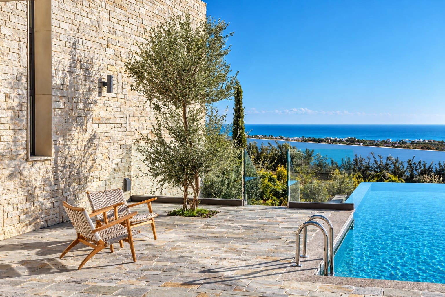 Stone-paved outdoor terrace with two armchairs and an olive tree beside the overflow swimming pool, overlooking the Mediterranean Sea