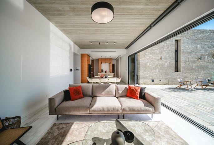 Furnished open-plan living area with sofa and coffee table, sliding glass doors opening onto a stone terrace, with dining and kitchen area beyond